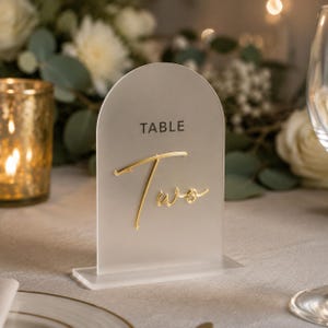 May include: An arched, frosted acrylic table sign featuring the word TABLE in a dark sans-serif font and the word Two in a raised, gold-colored script. The sign is mounted on a rectangular, clear acrylic base for upright display.