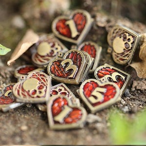 Puede incluir: Un conjunto de amuletos metálicos en forma de corazón con los números 1, 3 y 5 en dorado. Los amuletos tienen un centro rojo texturizado y un borde de filigrana dorada. Están esparcidos sobre tierra con hojas verdes.