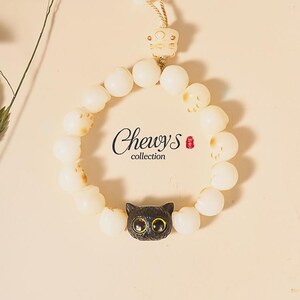 May include: A white beaded bracelet with a black cat charm. The bracelet features a cat-shaped charm with gold-coloured eyes and the text "Chewys collection". The bracelet is strung on a beige cord and has a cat-shaped clasp.