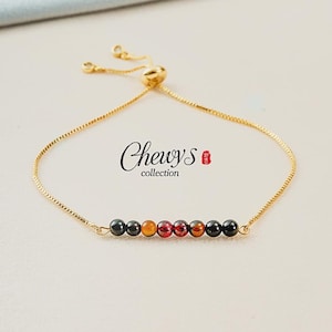 Pode incluir: Pulseira ajustável dourada com uma fileira de contas coloridas. As contas são pretas, laranja e vermelhas. A pulseira tem uma corrente fina e um fecho deslizante. O texto "Chewys collection" está impresso na imagem.