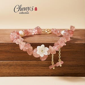 May include: A bracelet featuring pink strawberry quartz chip beads, white pearl accents, and a central white flower charm. Two gold-toned chains with dangling pink quartz chips hang from the flower. The text Chewy's collection is visible.