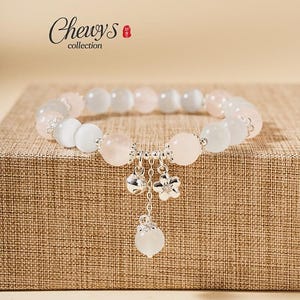 May include: A beaded bracelet with a mix of pink and white round beads. The bracelet features silver charms, including a flower and a teardrop-shaped bead. The brand name "Chewys collection" is visible in the upper left corner.