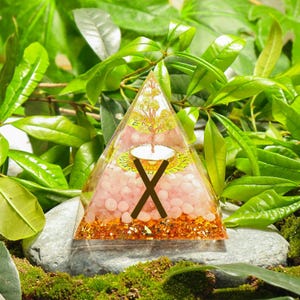 May include: A clear pyramid-shaped resin art piece with a gold base and a pink stone interior. A gold tree design is visible inside, along with a white and gold circular design and a brown 'X' shape. The pyramid sits on a gray stone, surrounded by green foliage.