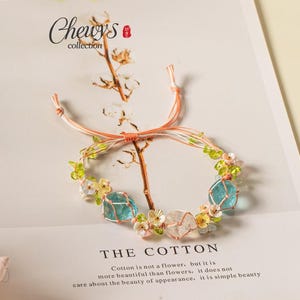 May include: A bracelet featuring light blue and clear raw stones, small green beads, and delicate flower-shaped charms strung on thin peach and white cord. The bracelet is displayed on a card with the text Chewy's collection and THE COTTON.