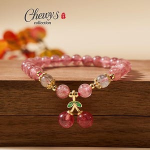 Op de afbeelding: Een roze kralenarmband met een kersenbedel. De armband heeft ronde, roze kralen en gouden accenten. De kersenbedel heeft twee rode, ronde kralen met groene blaadjes. De tekst "Chewy's collection" staat linksboven.