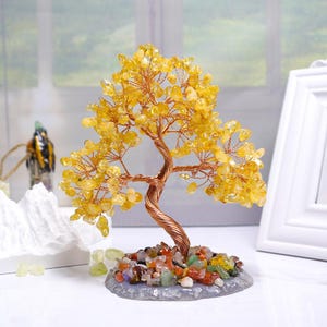 May include: A decorative tree sculpture with a copper wire trunk and branches, adorned with yellow gemstone leaves. The base is a mix of colorful stones. The tree is displayed on a white surface.