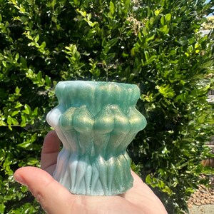 May include: A decorative candle holder with a unique, layered design. The candle holder features a gradient of colors, transitioning from white to light blue and then to a shimmering green with gold accents. The holder is held in a hand against a backdrop of green foliage.