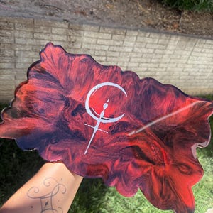 May include: A rectangular resin tray with an irregular, wavy edge featuring a marbled pattern of deep red and black swirls. A white graphic symbol consisting of a crescent moon shape atop a vertical line with a crossbar is centered on the surface.