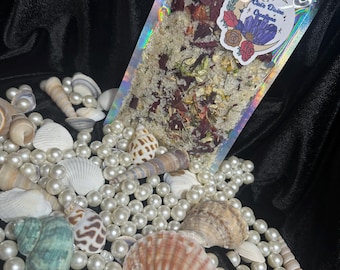 May include: A clear, iridescent bag filled with bath salts, dried flowers, and herbs. The bag has a sticker with the text "Gaia Divine Creations". The bag is surrounded by seashells, pearls, and a black velvet background.