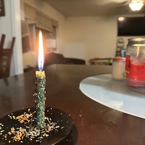 Może przedstawiać: Płonąca zielona świeca, ozdobiona ziołami, spoczywa na czarnym talerzu. Świeca stoi na ciemnobrązowym stole. Tło przedstawia rozmyty pokój.