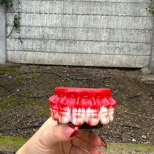May include: A red and white ceramic planter featuring multiple human faces around the exterior. The planter has a unique design with a dripping red paint effect. The planter is held in a hand.