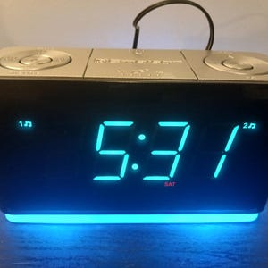 May include: A black and silver digital alarm clock displaying the time 5:31 AM on a Saturday. The clock has a blue LED display and two buttons on top. The base of the clock has a blue light.