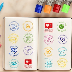 Pode incluir: Um caderno aberto com selos coloridos com frases como "Boa escrita", "Bom trabalho" e "Aprovado". Os selos são de várias cores, incluindo azul, verde e amarelo. Vários selos são visíveis na parte superior.