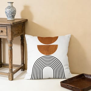 May include: White square throw pillow with a geometric design. The pillow features two brown half-circle shapes above a black and white striped arch on a white background. A vintage wooden table and a decorative vase are in the background.