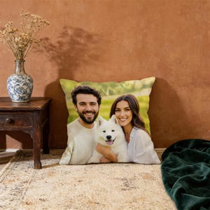 May include: A square throw pillow featuring a printed color photograph of a smiling man and woman posing outdoors with a fluffy white dog. The image on the pillow shows the three subjects centered against a blurred green background of grass and trees.