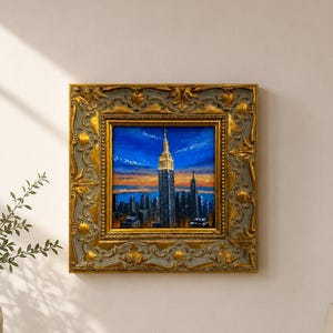 May include: A square oil painting depicting the Empire State Building at sunset, featuring a vibrant blue sky and orange horizon. The artwork is encased in an ornate, heavy gold-toned frame with intricate relief detailing and a distressed finish.