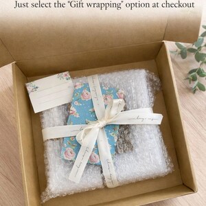May include: A brown cardboard box containing a rectangular item wrapped in clear bubble wrap, tied with a cream-colored ribbon and a blue floral patterned card. A small blank gift tag with floral accents is included. Text reads Gift Wrappable.