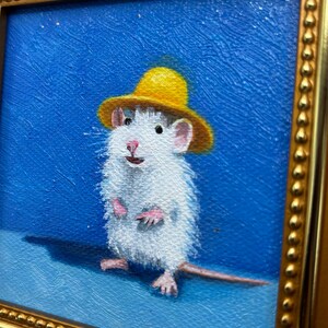 May include: An oil painting of a white rat wearing a small yellow hat against a solid blue background. The rat is standing upright with pink paws and a long tail. The artwork is enclosed in a decorative gold frame with a beaded edge detail.