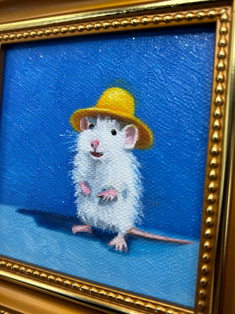May include: An oil painting of a white rat wearing a small yellow hat against a solid blue background. The rat is standing upright with pink paws and a long tail. The artwork is enclosed in a decorative gold frame with a beaded edge detail.