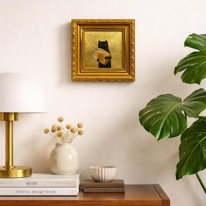 May include: A framed artwork featuring a black cat holding a round object, set against a gold background. The artwork is in a gold frame and hangs on a white wall. A lamp, vase, and books are on a wooden cabinet.