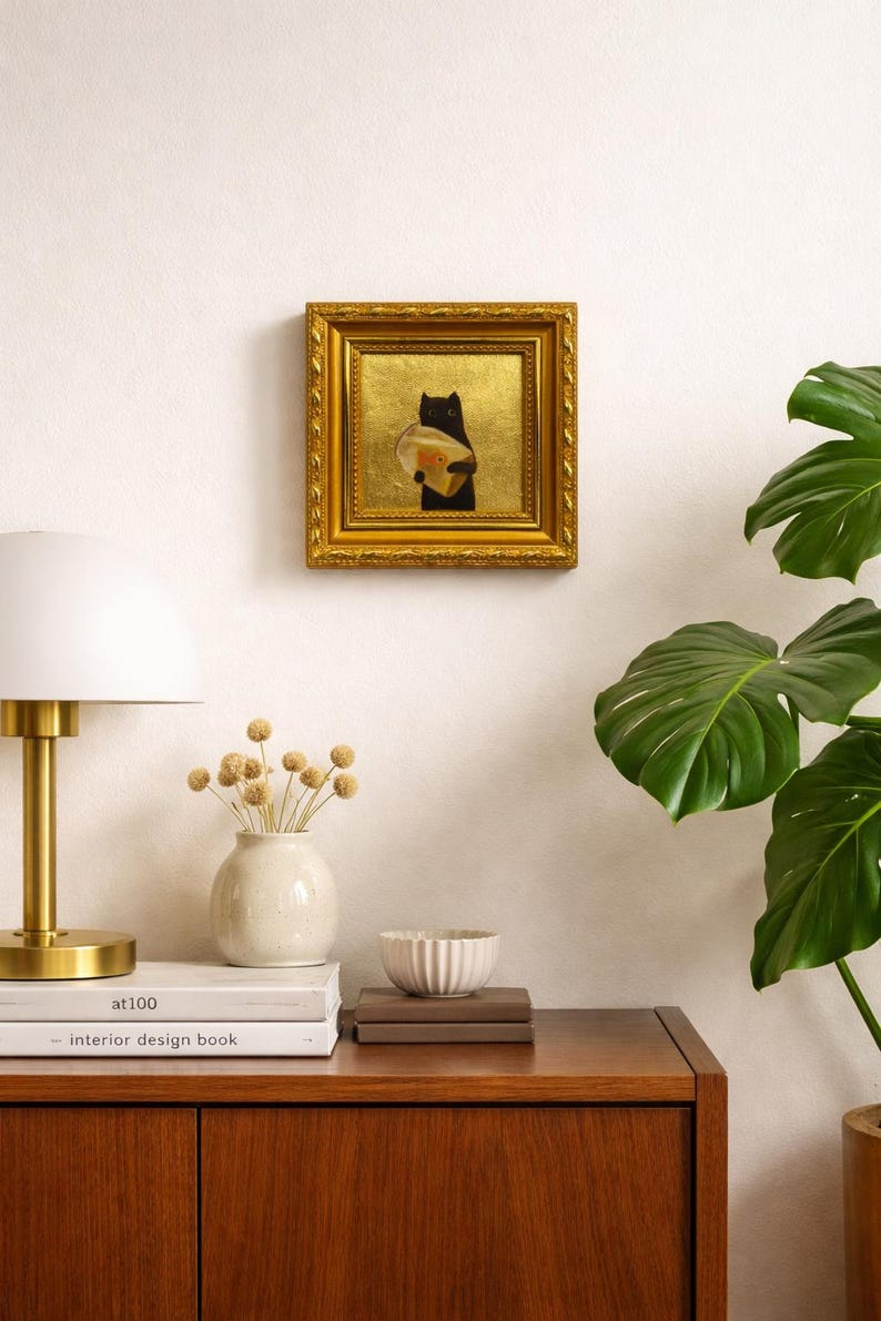 May include: A framed artwork featuring a black cat holding a round object, set against a gold background. The artwork is in a gold frame and hangs on a white wall. A lamp, vase, and books are on a wooden cabinet.