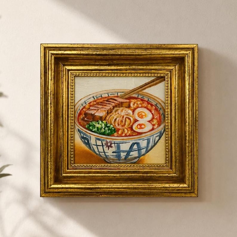 May include: A square painting of a bowl of ramen in a thick, ornate gold frame. The artwork depicts a blue and white patterned ceramic bowl filled with noodles, sliced pork, two soft-boiled egg halves, and green onions, with chopsticks.
