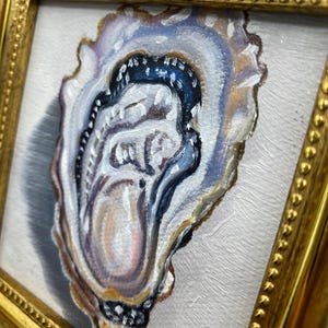 May include: Oil painting of an oyster shell in a gold frame. The shell is painted with shades of blue, white, and tan, with a dark blue interior. The frame has a beaded edge. The artwork is on a white background.