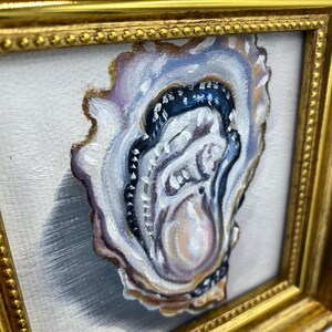 May include: An oyster painting in a gold frame. The oyster is painted with shades of white, purple, brown, and black. The painting is on a white background and is framed in a gold frame.