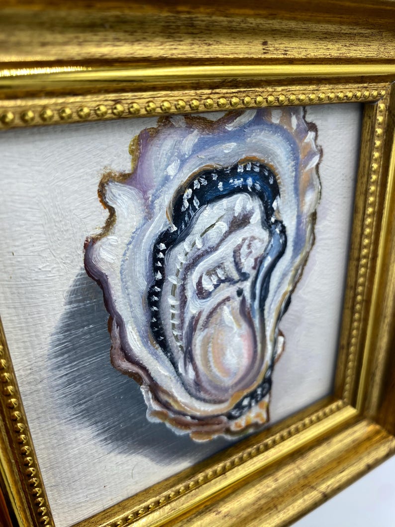 May include: An oyster painting in a gold frame. The oyster is painted with shades of white, purple, brown, and black. The painting is on a white background and is framed in a gold frame.