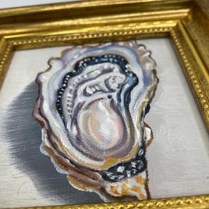 May include: A framed painting of an oyster shell. The artwork features a detailed depiction of the oyster's interior, with shades of white, blue, brown, and gold. The frame is a gold color.