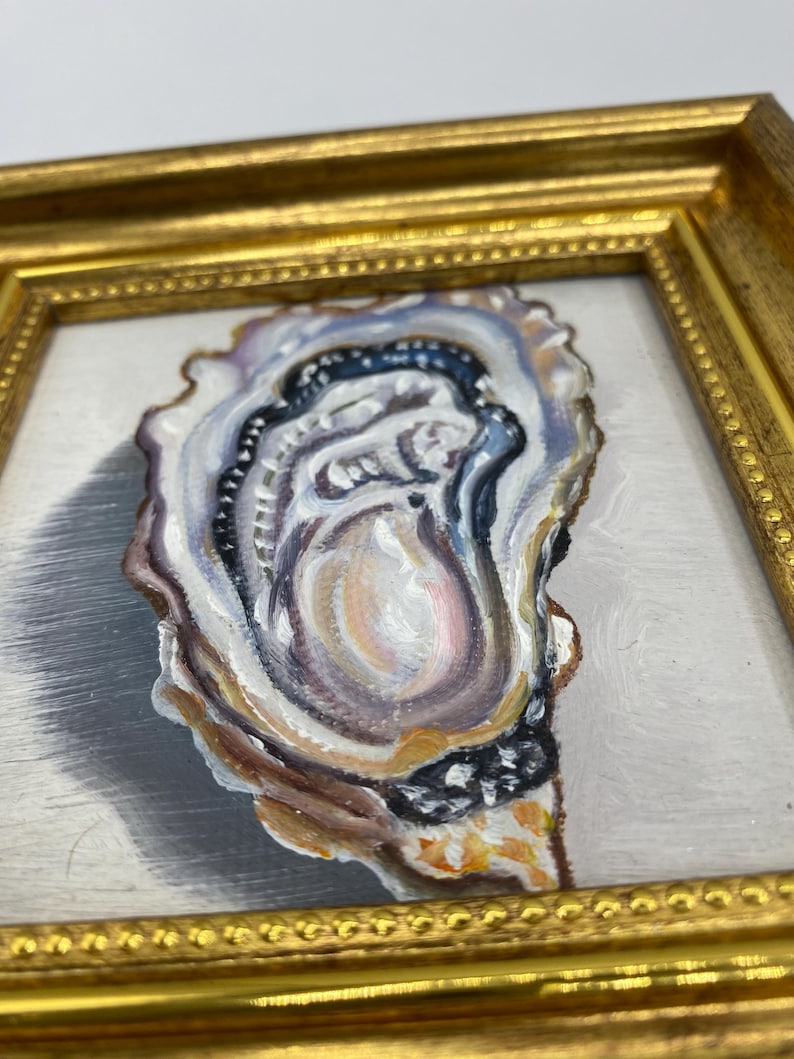 May include: A framed painting of an oyster shell. The artwork features a detailed depiction of the oyster's interior, with shades of white, blue, brown, and gold. The frame is a gold color.