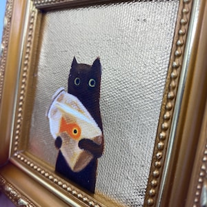 May include: A framed painting depicts a black cat holding a bag with a goldfish. The artwork is set against a gold textured background and framed in a gold ornate frame. The cat has large, round eyes.