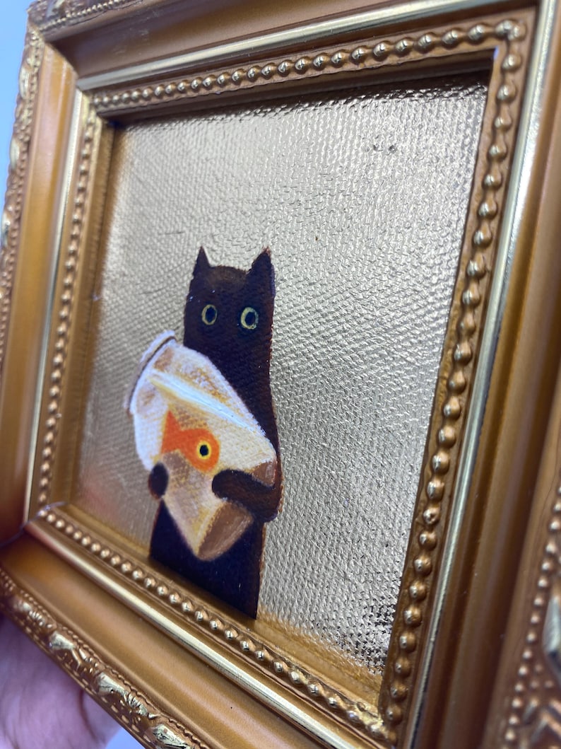 May include: A framed painting depicts a black cat holding a bag with a goldfish. The artwork is set against a gold textured background and framed in a gold ornate frame. The cat has large, round eyes.