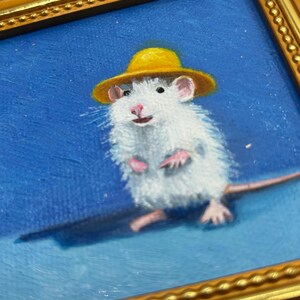 May include: An oil painting in a gold-toned ornate frame depicting a white mouse wearing a small yellow hat against a solid blue background. The mouse is shown in a standing position with pink paws and ears, rendered with visible brushstrokes.