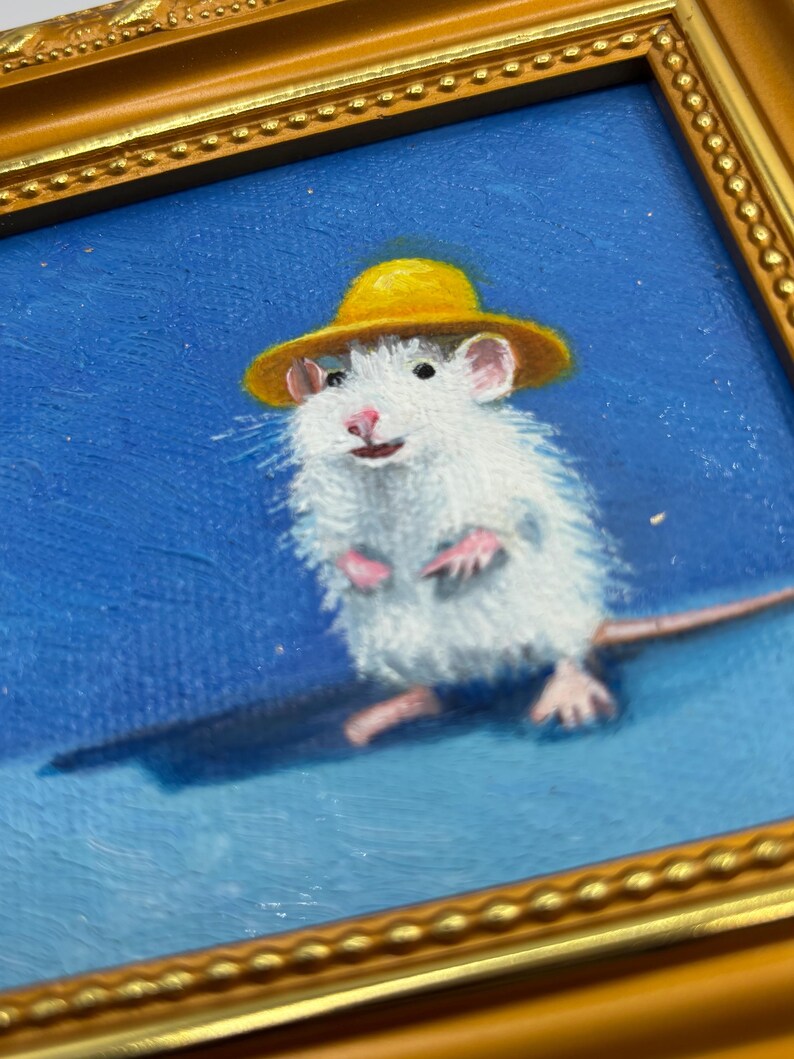 May include: An oil painting in a gold-toned ornate frame depicting a white mouse wearing a small yellow hat against a solid blue background. The mouse is shown in a standing position with pink paws and ears, rendered with visible brushstrokes.