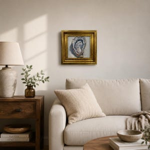 May include: A framed oyster painting with a gold ornate frame. The artwork features a detailed depiction of an oyster shell in shades of blue, white, and beige. The painting is displayed on a neutral-colored wall above a beige sofa.
