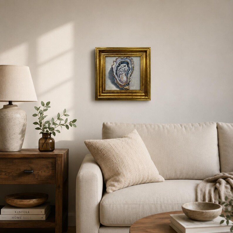 May include: A framed oyster painting with a gold ornate frame. The artwork features a detailed depiction of an oyster shell in shades of blue, white, and beige. The painting is displayed on a neutral-colored wall above a beige sofa.