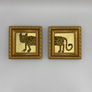 May include: Two framed paintings with gold leaf backgrounds and gold frames. Each painting features a stylized leopard with black spots. The leopard is depicted in a whimsical style, with a gold background.