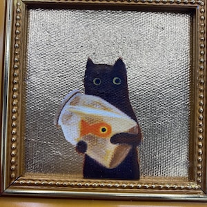May include: A framed painting of a black cat holding a fishbowl with a goldfish. The painting has a gold frame and a textured gold background. The cat has large, round eyes.