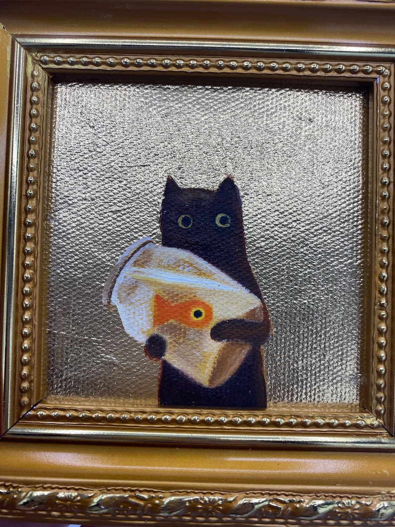 May include: A framed painting of a black cat holding a fishbowl with a goldfish. The painting has a gold frame and a textured gold background. The cat has large, round eyes.