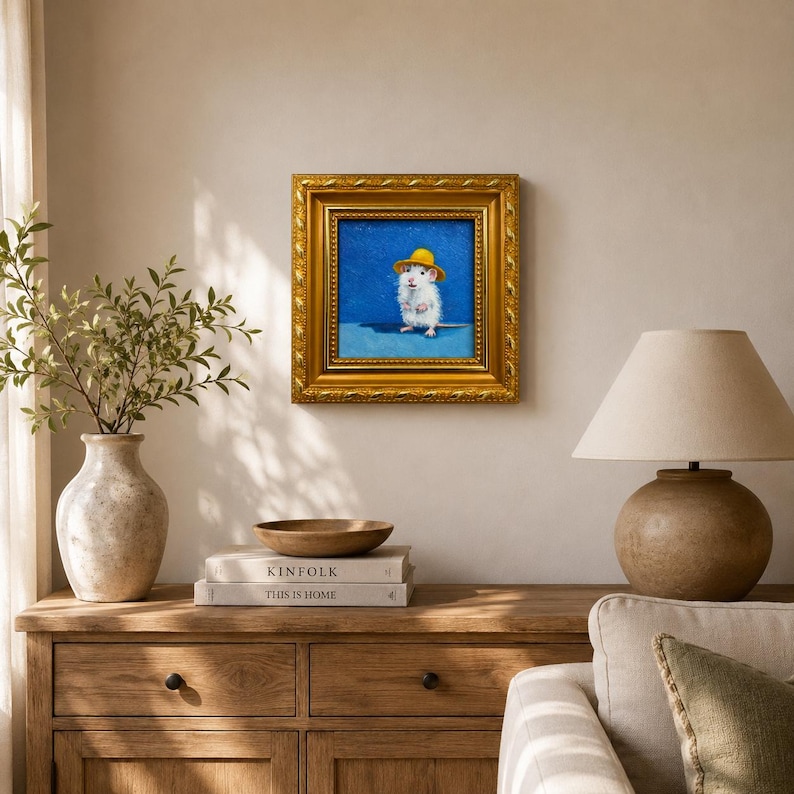 May include: A square painting in a decorative gold frame featuring a white mouse wearing a yellow hat against a solid blue background. The artwork is displayed on a wall, showcasing the detailed brushwork of the mouse and its bright yellow accessory.