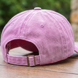 May include: A pink baseball cap with a washed-out fabric texture, featuring a six-panel construction with stitched eyelets, a top button, and an adjustable strap at the back secured with a rectangular bronze-toned metal sliding buckle.