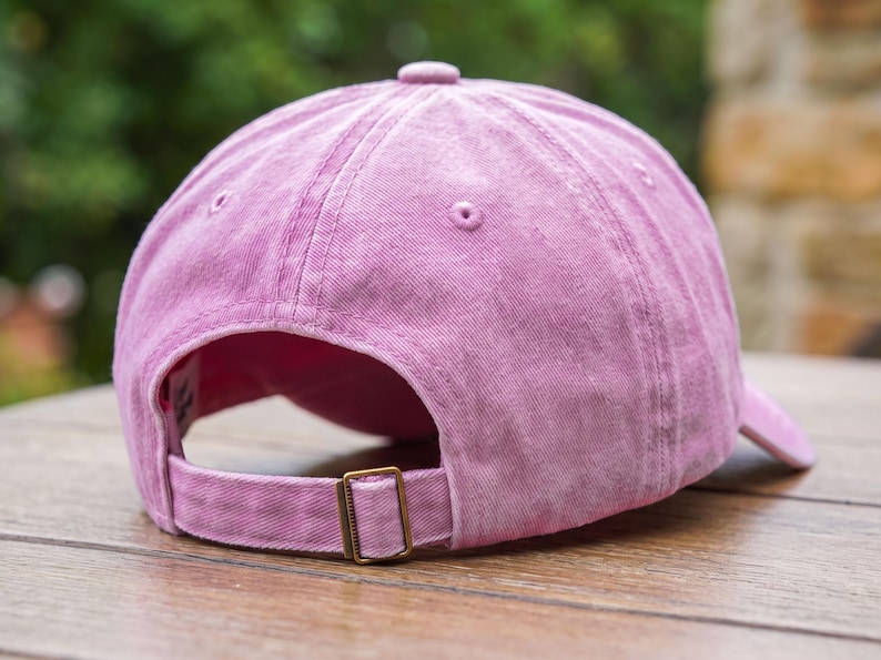 May include: A pink baseball cap with a washed-out fabric texture, featuring a six-panel construction with stitched eyelets, a top button, and an adjustable strap at the back secured with a rectangular bronze-toned metal sliding buckle.