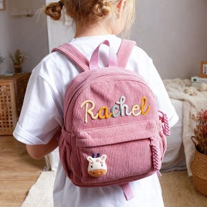 May include: A pink corduroy backpack with the name "Rachel" embroidered in colorful letters. The backpack has a small front pocket and a giraffe patch. It features adjustable straps and a top handle.