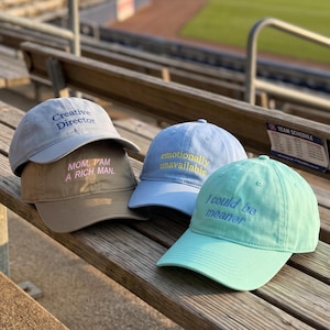Op de afbeelding: Vier baseballpetten in lichtblauw, beige, maagdenpalm en mintgroen. De maagdenpalmkleurige pet zegt Creative Director, de beige pet zegt MOM, I AM A RICH MAN., de lichtblauwe pet zegt emotionally unavailable en de mintgroene pet zegt I could be meaner.