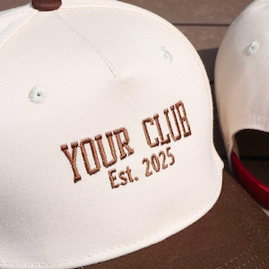 May include: Cream-colored baseball cap with a brown brim and top button. The words "YOUR CLUB Est. 2025" are embroidered in brown thread on the front. A second cap is visible in the background.