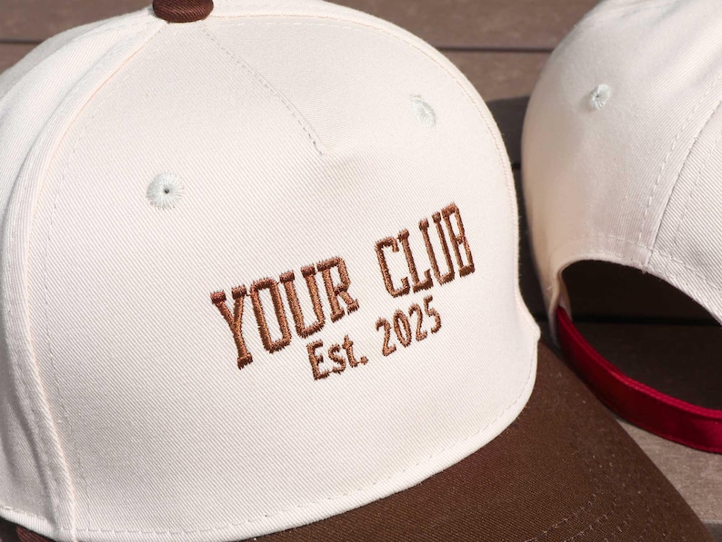 May include: Cream-colored baseball cap with a brown brim and top button. The words "YOUR CLUB Est. 2025" are embroidered in brown thread on the front. A second cap is visible in the background.