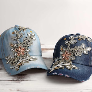 Puede incluir: Se muestran dos gorras de béisbol de mezclilla, una azul claro y otra azul oscuro. Ambas gorras presentan elaborados adornos florales con cristales y cuentas transparentes y color melocotón. Las gorras están sobre un fondo de madera blanca.