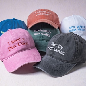 May include: A collection of six baseball caps in various colors including pink, blue, terracotta, dark green, white, and charcoal gray. Each cap features embroidered text such as I need a Diet Coke, I could be meaner, emotionally unavailable, and Heavily Caffeinated.