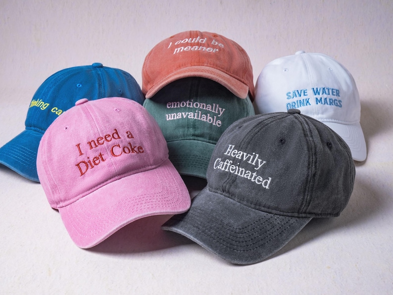 May include: A collection of six baseball caps in various colors including pink, blue, terracotta, dark green, white, and charcoal gray. Each cap features embroidered text such as I need a Diet Coke, I could be meaner, emotionally unavailable, and Heavily Caffeinated.